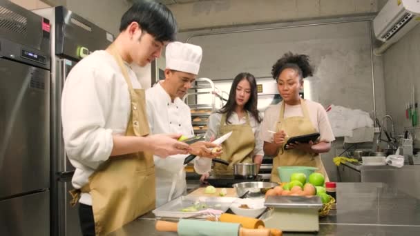 趣味の料理コース 料理の制服を着た上級男性シェフは 若い料理クラスの学生にリンゴの皮をむいて切ること ペストリー料理の材料 レストランのステンレススチールキッチンのフルーツパイを教えています — ストック動画