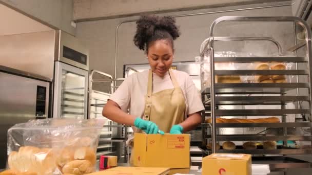ベーカリースタートアップ小規模なビジネス配信 アフリカ系アメリカ人女性の料理人の一人は 手作りの焼きたてのパンやペストリーを箱に詰め 料理キッチンでオンライン顧客の購入のために送っています — ストック動画