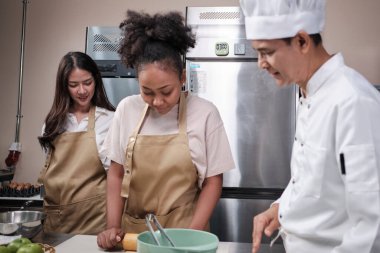 Mutfak kursu, aşçı üniformalı kıdemli erkek aşçı genç aşçılık sınıfı öğrencilerine hamur yoğurmayı ve rulo yapmayı öğretiyor, fırın yemekleri için malzemeler hazırlıyor, paslanmaz çelik mutfakta meyveli turtalar pişiriyor..