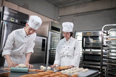 Beyaz üniformalı ve önlüklü iki profesyonel Asyalı aşçı hamur ve yumurta yoğurur, ekmek, kurabiye ve taze fırın yemeği hazırlar, paslanmaz çelik bir mutfakta fırında pişirir..