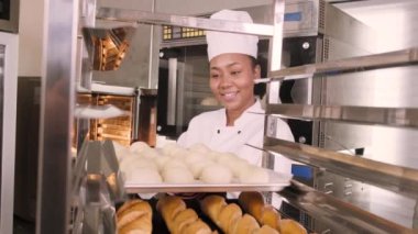 Profesyonel Afro-Amerikan kadın aşçı üniforması, eldiven ve önlük, hamur işiyle ekmek yapmak, taze fırın yemekleri hazırlamak, restoranın paslanmaz çelik mutfağında fırında pişirmek..