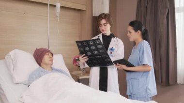 Üniformalı beyaz kadın doktor, Asyalı radyologla röntgen filmini ve iyileşen erkek hastayı hastane koğuşunda yatağında, tıbbi klinikte ve kanser muayenesinde açıklıyor..