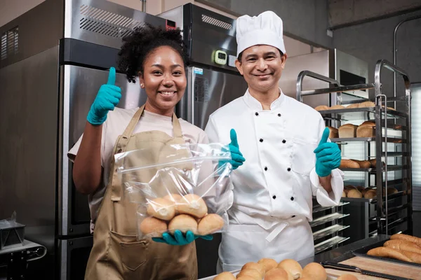 Üniformalı iki profesyonel şefin portresi. Neşeli, gururlu bir gülümsemeyle kameraya bakıyor ve mutfakta ekmeği parmaklarıyla kaldırıyor. Ticari hamur işlerinin ve taze fırın mutfağının ortağı..