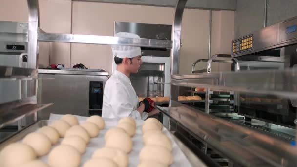ペストリー生地からパンを作る帽子 エプロンで統一された白いコックの若いプロのアジアの男性シェフ 新鮮なベーカリーフードを準備し レストランのステンレススチールキッチンでオーブンで焼く — ストック動画