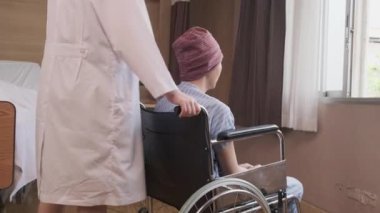 Üniformalı beyaz kadın terapist tekerlekli sandalyeli hastayı pencereye itiyor, kemoterapi sonrası kanser hastalığının iyileşmesini destekliyor ve teşvik ediyor..