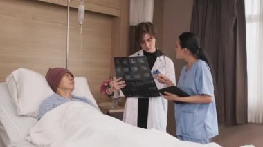 Üniformalı beyaz kadın doktor, Asyalı radyologla röntgen filmini ve iyileşen erkek hastayı hastane koğuşunda yatağında, tıbbi klinikte ve kanser muayenesinde açıklıyor..