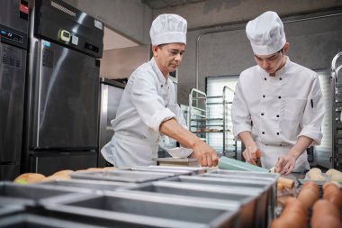 Beyaz aşçı üniforması ve önlükleri içinde iki profesyonel Asyalı şef hamur ve yumurta yoğuruyor, ekmek ve taze fırın yemekleri hazırlıyor, restoranın paslanmaz çelik mutfağında fırında pişiriyorlar..