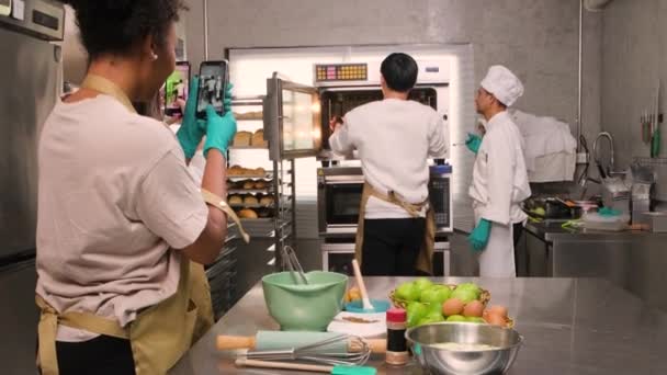 年轻的亚洲男性烹饪班学生从电炉中拿来一盘烤馅饼 旁边是资深厨师 在烹饪课上教糕点烹饪 在不锈钢厨房里教食物 — 图库视频影像