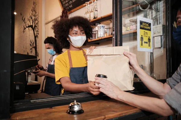 Yüz maskeli Afro-Amerikalı kadın barista sosyal mesafeli kafede çalışıyor, müşteriye kahve ısmarlıyor COVID19 karantina altındaki küçük işletme kahve dükkanının yeni normal hizmeti..