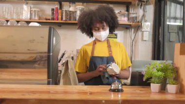 Afro-Amerikalı bayan barista kahve fincanlarını temizler, kafenin camından bakar, yeni normal yaşam tarzı servisinde müşterileri bekler, COVID19 pandemik karantinasının KOBİ iş etkisi.