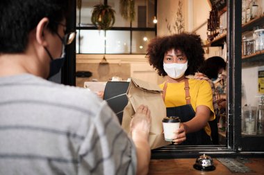 Yüz maskeli Afro-Amerikalı kadın barista sosyal mesafeli kafede çalışıyor, müşteriye kahve ısmarlıyor COVID19 karantina altındaki küçük işletme kahve dükkanının yeni normal hizmeti..