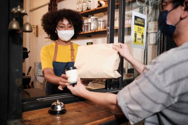 Yüz maskeli Afro-Amerikalı kadın barista sosyal mesafeli kafede çalışıyor, müşteriye kahve ısmarlıyor COVID19 karantina altındaki küçük işletme kahve dükkanının yeni normal hizmeti..