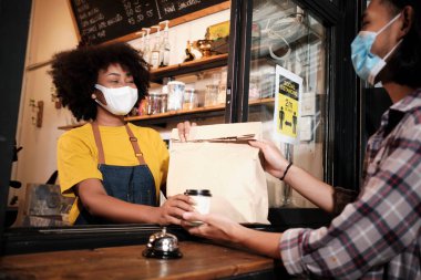 Yüz maskeli Afro-Amerikalı kadın barista sosyal mesafeli kafede çalışıyor, müşteriye kahve ısmarlıyor COVID19 karantina altındaki küçük işletme kahve dükkanının yeni normal hizmeti..
