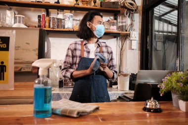 Bir kafede sağlıklı korunma için alkol dezenfektanı, yüz maskeli Asyalı erkek barista dizüstü bilgisayar kullanıyor, COVID19 pandemik karantinasında kahve siparişini bekliyor, bu da küçük işletmeler için ekonomik etki..