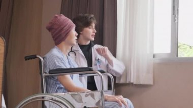 Uniformed young Caucasian female therapy doctor encouraging wheelchair male patient at window to support and motivate recovery, cancer illness after chemo medical treatment in hospital inpatient room.