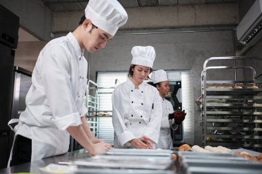 Profesyonel gurme takımı, beyaz aşçı üniforması ve önlüklü üç genç aşçı hamur ve yumurta yoğurur, ekmek ve taze fırın yemeği pişirir paslanmaz çelik restoran mutfağında fırında pişirir..