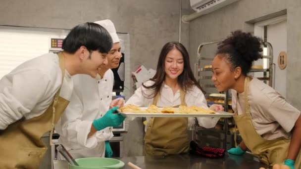 若いアジアの女性料理クラスの学生は オーブンから焼きパイのトレイを示しています シェフのグループのための良い香り 料理コースの授業で幸せなペストリー料理 ステンレス製のキッチンでの食品職業 — ストック動画