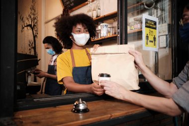 Yüz maskeli Afro-Amerikalı kadın barista sosyal mesafeli kafede çalışıyor, müşteriye kahve ısmarlıyor COVID19 karantina altındaki küçük işletme kahve dükkanının yeni normal hizmeti..