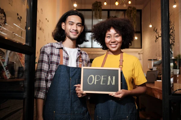 Two young startup barista partners with aprons stand at casual cafe door, letters on board and show open sign, happy and cheerful smiles with coffee shop service jobs, and new business entrepreneurs.