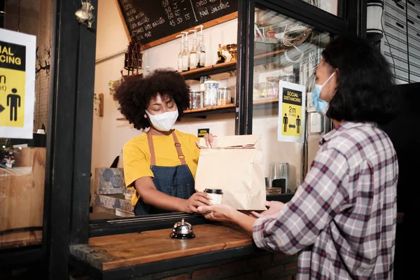 Yüz maskeli Afro-Amerikalı kadın barista sosyal mesafeli kafede çalışıyor, müşteriye kahve ısmarlıyor COVID19 karantina altındaki küçük işletme kahve dükkanının yeni normal hizmeti..