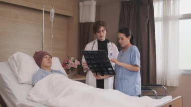 Üniformalı beyaz kadın doktor, Asyalı radyologla röntgen filmini ve iyileşen erkek hastayı hastane koğuşunda yatağında, tıbbi klinikte ve kanser muayenesinde açıklıyor..