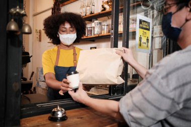 Yüz maskeli Afro-Amerikalı kadın barista sosyal mesafeli kafede çalışıyor, müşteriye kahve ısmarlıyor COVID19 karantina altındaki küçük işletme kahve dükkanının yeni normal hizmeti..