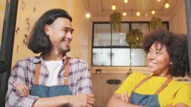 Two young startup barista partners with aprons stand at casual cafe door, arms crossed, laugh and tease together, happy and cheerful smiles with coffee shop service jobs, small business entrepreneurs.