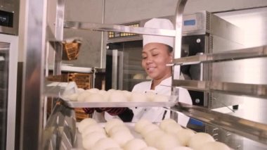 Profesyonel Afro-Amerikan kadın aşçı üniforması, eldiven ve önlük, hamur işiyle ekmek yapmak, taze fırın yemekleri hazırlamak, restoranın paslanmaz çelik mutfağında fırında pişirmek..