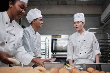 Profesyonel gurme takımı, beyaz aşçı üniforması ve önlüklü üç şef hamur ve yumurta yoğurur, ekmek ve taze fırın yemeği pişirir paslanmaz çelik restoranda fırında pişirir..