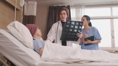 Üniformalı beyaz kadın doktor, Asyalı radyologla röntgen filmini ve iyileşen erkek hastayı hastane koğuşunda yatağında, tıbbi klinikte ve kanser muayenesinde açıklıyor..