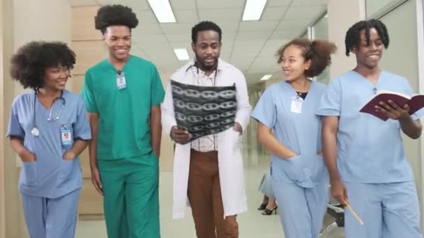 专业的非洲裔美国男性医生与医科学生一起步行 在泰国的诊所医院讨论诊断用的X光片 墙上的文字表示药物和财务 — 图库视频影像
