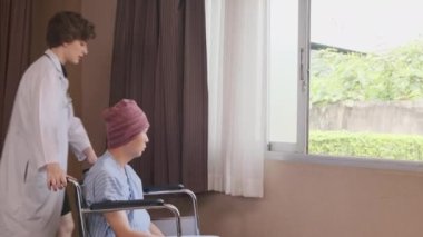 Uniformed young Caucasian female therapy doctor pushes wheelchair male patient to window, supports and motivates recovery from cancer illness after chemo medical treatment in hospital inpatient room.