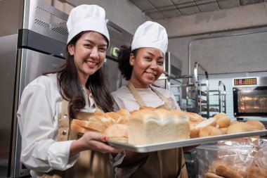 İki aşçı üniformalı genç kadın aşçının portresi. Neşeli bir gülümsemeyle kameraya bakıyor ve mutfakta bir tepsi ekmekle gururlanıyor. Pastanenin arkadaşı ve ortağı, günlük taze fırın işi..