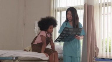 Asian female doctor in uniform health checks African American child, illness patient, diagnosis explains x-ray film in emergency room bed at hospital ward, pediatric clinic, kids examination consult.