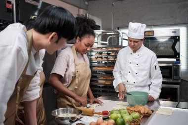 Mutfak kursu, aşçı üniformalı kıdemli erkek aşçı genç aşçılık sınıfı öğrencilerine hamur yoğurmayı ve rulo yapmayı öğretiyor, fırın yemekleri için malzemeler hazırlıyor, paslanmaz çelik mutfakta meyveli turtalar pişiriyor..