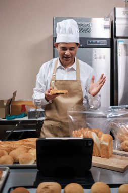Önlüklü kıdemli Asyalı erkek şef, küçük işletme blogcusu mobil uygulama teknolojisi, fırın satışları online promosyon, taze ekmek, hamur işi ve mutfakta ticari aşçılık yoluyla canlı yayın yapıyor..