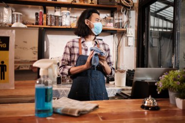 Yüz maskeli Asyalı erkek barista dizüstü bilgisayar kullanıyor, COVID19 pandemik karantinasında kahve siparişini bekliyor, kafede sağlıklı korunma için alkol dezenfektanını kullanıyor, bunun küçük işletmeler için ekonomik etkisi.