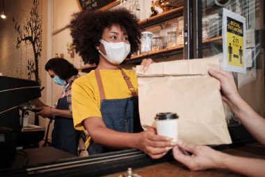 Yüz maskeli Afro-Amerikalı kadın barista sosyal mesafeli kafede çalışıyor, müşteriye kahve ısmarlıyor COVID19 karantina altındaki küçük işletme kahve dükkanının yeni normal hizmeti..