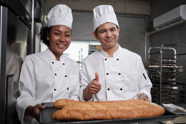 Beyaz üniformalı profesyonel şeflerin portresi kameraya neşeli bir gülümsemeyle bakıyor ve mutfakta bir tepsi baget ekmeğiyle gurur duyuyor. Pasta yiyecekleri ve taze günlük fırın işlerinin arkadaşı ve ortağı..
