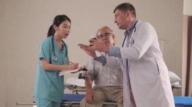 Profesyonel Kafkas erkek doktoru yaşlı hastaya tavsiye eder ve hastane koğuşundaki kadın tıp öğrencilerine reçeteli ilaçları, hastalık danışmanlıklarını ve sağlık muayenelerini açıklar..