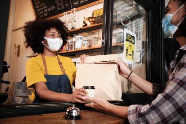 Yüz maskeli Afro-Amerikalı kadın barista sosyal mesafeli kafede çalışıyor, müşteriye kahve ısmarlıyor COVID19 karantina altındaki küçük işletme kahve dükkanının yeni normal hizmeti..
