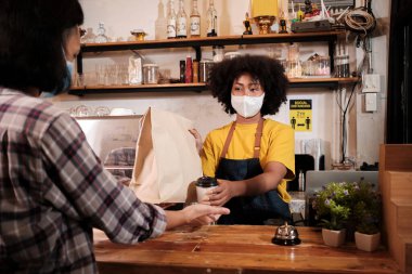 Yüz maskeli Afro-Amerikalı kadın barista sosyal mesafeli kafede çalışıyor, müşteriye kahve ısmarlıyor COVID19 karantina altındaki küçük işletme kahve dükkanının yeni normal hizmeti..