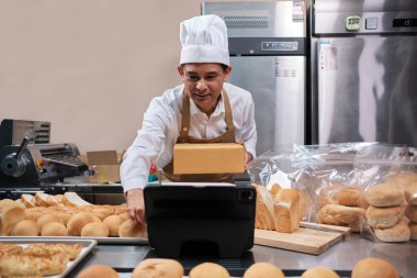 Önlüklü kıdemli Asyalı erkek şef, küçük işletme blogcusu mobil uygulama teknolojisi, fırın satışları online promosyon, taze ekmek, hamur işi ve mutfakta ticari aşçılık yoluyla canlı yayın yapıyor..