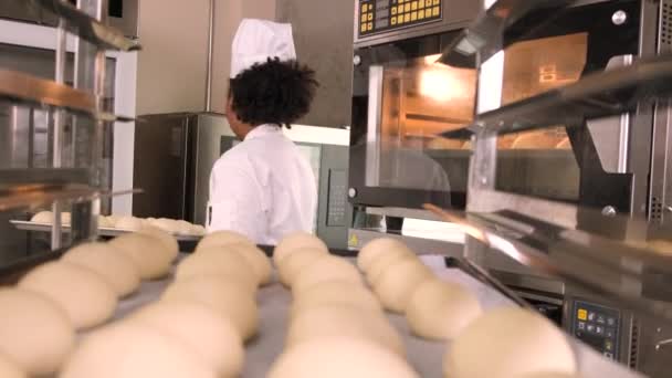 身穿白色厨师制服 手套和围裙的非洲裔美国女厨师 用糕点面团做面包 准备新鲜的烘焙食品 在餐馆不锈钢厨房的烤箱里烘焙 — 图库视频影像