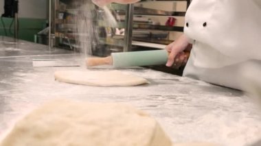 Yavaş çekimde ve yakın planda, önlüklü beyaz aşçı üniformalı şef un serpiyor, hamur işlerini rulo ile yoğuruyor, ekmek, turta ve restoranın paslanmaz çelik mutfağında taze fırın yemekleri hazırlıyor..