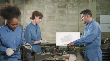 Üç çok ırklı profesyonel endüstri mühendisi güvenlik üniformalı işçiler metal işçiliği işleri bir monitör, torna makineleri ve imalat fabrikasında atölye ile mekanik çizim tartışır.
