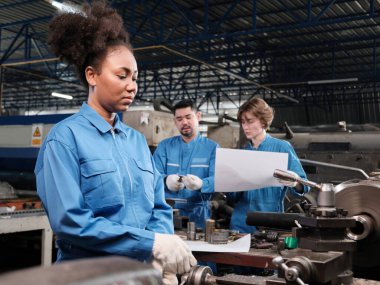 Profesyonel bir Afrikalı Amerikalı kadın mühendis güvenlik üniforması içinde çalışıyor. Metal işçiliği hassas aletleri, mekanik torna makineleri ve bir fabrikada yedek parça atölyesi var..
