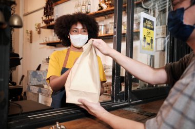 Yüz maskeli Afro-Amerikalı kadın barista sosyal mesafeli kafede çalışıyor, müşteriye kahve ısmarlıyor COVID19 karantina altındaki küçük işletme kahve dükkanının yeni normal hizmeti..