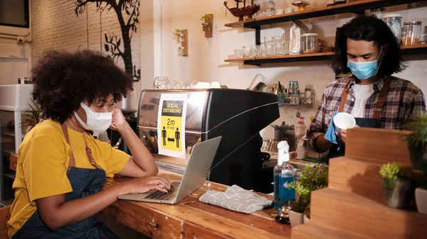 Two young cafe barista partners and entrepreneur work with face mask in coffee shop, waiting for customers order in new normal lifestyle service, SME business impact from COVID19 pandemic quarantine.