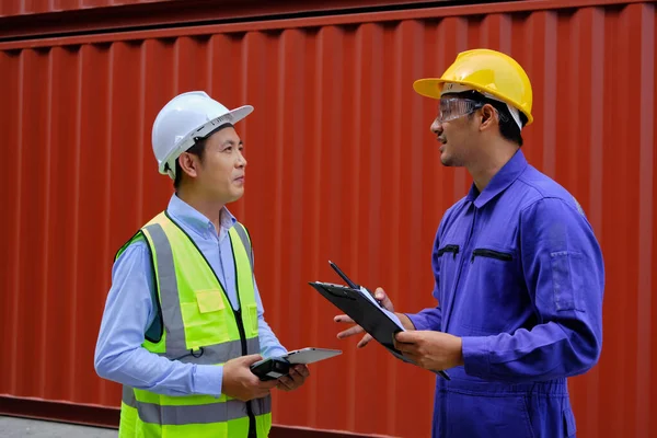 Two professional Asian male workers in safety uniforms and hard hats on a steel container background, work at logistics terminal, loading control shipping goods for the cargo transportation industry.